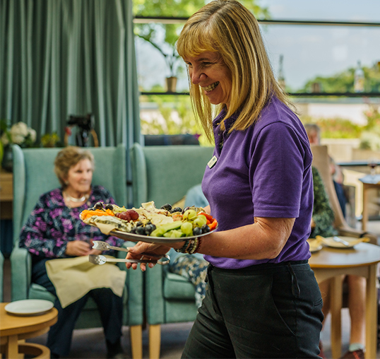 Image shows female member of staff serving a beautiful platter of cheese. She looks friendly and happy. 